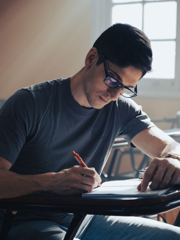 Serviços Young man focused on writing an exam in a classroom setting with natural light.