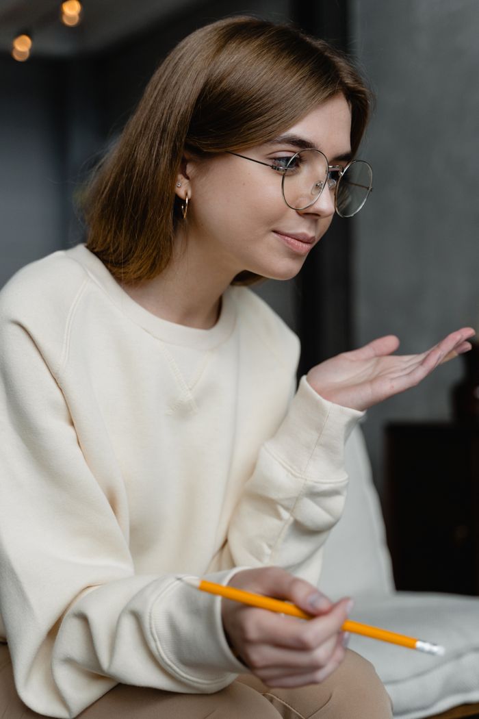 Serviços A woman in eyeglasses holding a pencil sits on a couch, engaged in thought.