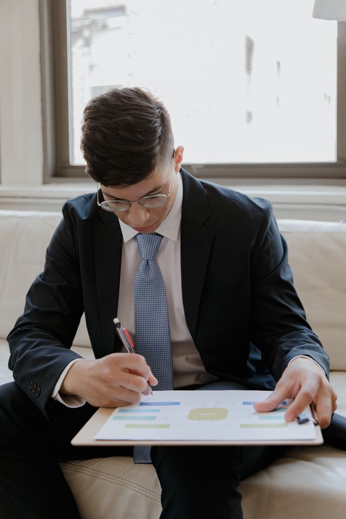 Serviços Young businessman in a suit writing on a clipboard while sitting indoors near a window.